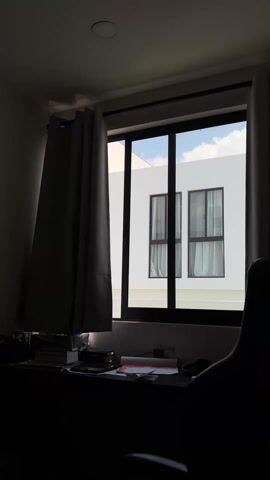 The scene presents a dimly lit room, with the primary focus on a window. The window, divided into six panes, reveals a view of a white building with two similar windows, also divided into panes, visible across from the room. The sky is visible through the panes, showing a blue sky with some white clouds. The window in the room is framed by gray curtains, with one curtain partially drawn to the left. In front of the window is a desk covered with books, papers, and other items. The room is generally dark, with the light primarily coming through the window, illuminating the exterior building. A portion of a chair is visible in the lower right corner of the frame. The overall impression is that of a workspace in a room with controlled lighting.