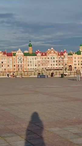 The video shows a wide outdoor plaza with a cobblestone surface. In the background, a row of colorful buildings with Dutch-style gables and red and green roofs stretch across the frame. A body of water, possibly a river or canal, runs in front of the buildings, separated by a black metal fence. Several people are visible in the plaza. Two women walk towards the camera on the right side of the plaza. Further in the background, several individuals are seated on benches, and some are standing near the fence. A lamppost stands near the center of the plaza. A tall, modern orange building with green accents is visible to the left of the colorful buildings. The sky is cloudy with patches of blue. A shadow of a person is cast on the ground in the foreground, indicating the presence of the videographer. The video is a static shot.