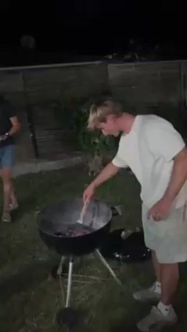Here is a description of the video:

The video shows a person tending to a charcoal grill at night. The person is wearing a white t-shirt and beige shorts. The grill is a round, black kettle-style grill. In the background, there is a wooden fence and some shrubbery.  Another person is standing off to the side and is partially visible. The camera then pans over to show a swimming pool and focuses on a woman with brown curly hair wearing large glasses, who gives a thumbs up and smiles.