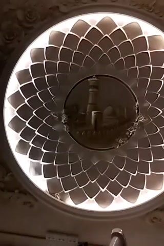 The video shows a close-up, low-angle shot of an ornate ceiling light fixture. The fixture is circular and features a central medallion with a relief carving of a minaret and a dome, likely a representation of a mosque. Surrounding the medallion is a radial pattern of interlocking, petal-like shapes that are backlit, creating a glowing effect. The overall aesthetic suggests a traditional or classical architectural style. The lighting is dim, with the primary illumination coming from the fixture itself. No people or other significant objects are visible in the frame.