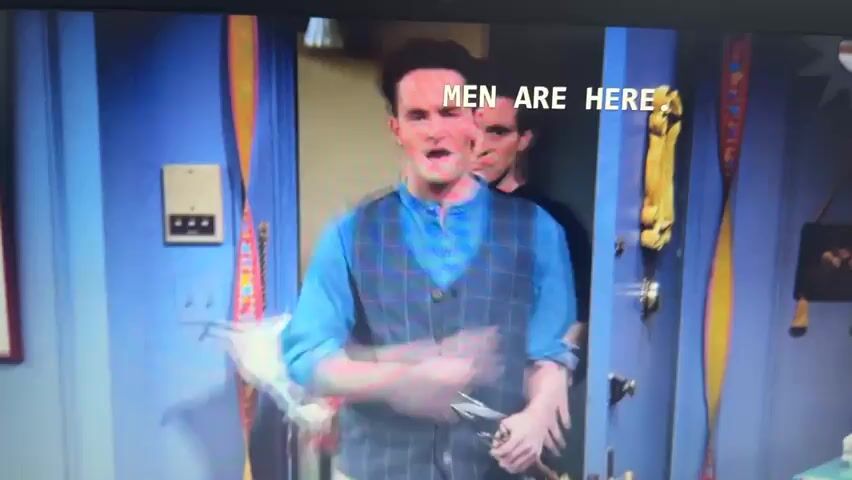 Two men stand side by side in a hallway. The man on the left, wearing a black t-shirt, holds a package. The man on the right, wearing a blue shirt and a plaid vest, holds grilling tongs and a barbecue lighter. Text overlayed on the image reads "MEN ARE HERE." The scene then shifts to two women standing in a room decorated with balloons and flowers. The woman on the left has blonde hair and is smiling, while the woman on the right has dark hair and a serious expression. The scene then returns to the two men.