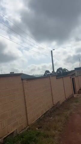 This media file shows a view of a street in Gembu, Nigeria, with a brick wall and a dirt road. The sky is overcast with dark clouds, suggesting it is either early morning or late afternoon, or possibly just before a storm. The wall is made of light brown bricks and has a green coping. There are power lines running above the wall and a streetlight on a pole. A person in a dark coat and a hat is walking away from the camera down the dirt road. Further down the road, two other figures are visible, also walking away. The background shows trees and buildings on a hillside. The overall mood is somewhat somber due to the cloudy sky and the muted colors of the scene. There is no visible text in the media file.