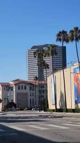 This is a vertical, first-person perspective video. The camera is moving and panning across a street scene under a clear blue sky. In the foreground, a crosswalk is visible. On the right side of the street, a large, light-colored building is prominent, featuring large billboards on its facade. One billboard displays a graphic of a character with a red cape and a yellow symbol. Another billboard features the words "JENNIFER HUDSON". The Warner Bros. logo and "GATE 2" are visible on the building. Palm trees are scattered throughout the scene, some tall and thin, others shorter. A white sedan drives across the intersection from left to right in front of the building. A tall, modern, glass-windowed building is visible in the background behind some of the palm trees and a lower, older building. An American flag and a California state flag are flying on poles atop the building. Traffic lights are visible at the intersection. The overall impression is an outdoor urban or studio lot setting on a sunny day.