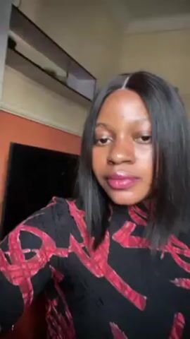 A young woman with straight black hair is facing the camera. She's wearing a black, long-sleeved top with a red abstract pattern. The background includes a cream-colored wall, white crown molding, an orange accent wall, a black television, and a white shelf. The woman appears to be talking, as her lips are moving.