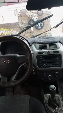 The video shows the interior of a Chevrolet Cobalt car. The steering wheel is visible on the left, and the dashboard is in the center of the frame. The gear stick is visible in the lower right corner. The windshield wipers are moving. Outside the windshield, there is a brick wall with several car wheel covers mounted on it. The car's interior appears clean and well-maintained. The video is shot from the driver's perspective.