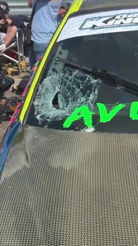 The video pans across a sunny outdoor event with several people and modified cars. The primary focus is on a blue and teal BMW with a damaged windshield, a carbon fiber hood, and the text "AVON FAB" written in green across the windshield. Nearby, another car, mostly green with a large graphic on the side, is parked. Several people are visible in the background, some standing, some sitting, near what appears to be a race track barrier and tents. The ground is paved, and the sky is blue with scattered clouds.