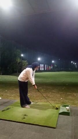 A person wearing a hijab and a white long-sleeved shirt over a striped shirt, and dark pants, is swinging a golf club at a golf driving range at night. The person is standing on a green mat. There are golf balls in a green holder to the right of the person. The driving range is lit by artificial lights. There are flags visible in the background. The person is mid-swing in the first frame and then has completed the swing in the subsequent frames.