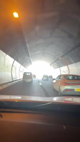 The video begins inside a car as it exits a tunnel. The camera is positioned from the driver's perspective. The tunnel is illuminated by bright light at the exit. Several cars are visible ahead, moving away from the tunnel. The tunnel walls are concrete and show some signs of wear. As the car emerges, the scene transitions to a highway under a bright blue sky with scattered clouds. The highway has multiple lanes, and other vehicles are driving on it. On the right side of the highway, there is a steep, rocky embankment with some cypress trees. On the left side, there is a guardrail and some infrastructure, including power lines and poles. The speed limit sign visible indicates 100. The overall environment is sunny and clear.