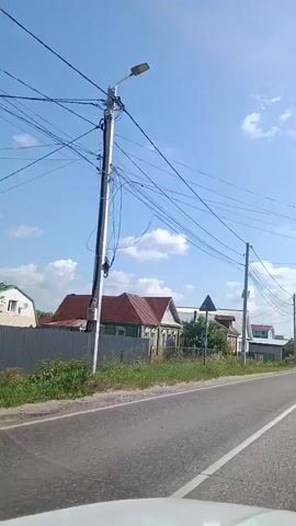 The video shows a view from a vehicle traveling on a paved road. The road is bordered by a grassy shoulder and some trees. On the left side of the road, there are several houses with red and green roofs, some with white fences. There are also power lines running overhead. The sky is blue with some white clouds. The video is from a first-person perspective, as if the viewer is in the car. The vehicle appears to be moving at a moderate speed.