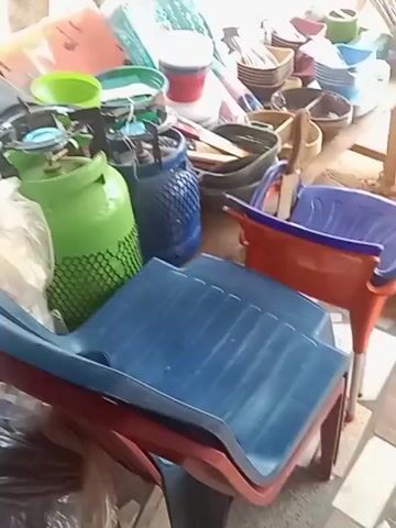 The video shows a variety of household items seemingly for sale. There are stacks of colorful plastic chairs, a green gas cylinder, plastic buckets, bowls, and containers. Some items are wrapped in plastic. Additionally, a box and some metal trays are visible. The camera pans slightly, revealing a wrapped item with a sticker and a blue plastic chair. The video appears to be taken in a shop or storage area.