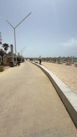 The video shows a wide, sandy beach with rows of straw umbrellas and empty lounge chairs. The sky is clear and bright blue, suggesting a sunny day. There are a few people scattered on the beach, some sitting under umbrellas and others walking. The foreground features a paved walkway next to a concrete barrier, separating it from the sand. In the distance, the ocean is visible with a clear blue horizon. A few bicycles are parked near a building on the left side of the frame. The overall impression is of a calm, well-maintained beach resort.