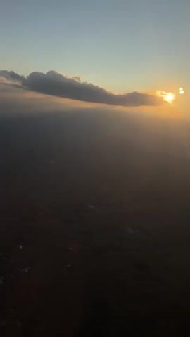 The video is a first-person view from an airplane window, looking down at the landscape below. The sun is visible in the upper right corner, partially obscured by a cloud. The sun is setting, casting a warm orange glow across the sky. The landscape below is dark and appears to be farmland with some scattered buildings. There are some faint watermarks or smudges on the window, visible as streaks and spots, particularly in the lower right portion of the frame. The overall impression is one of a peaceful descent at dusk.