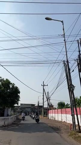 The video shows a street scene with several motorbikes riding down a paved road under a clear blue sky. The road is lined with buildings on the left and a wall painted with red and white stripes on the right. A prominent feature of the scene is a dense network of overhead electrical wires, supported by poles, which create a complex, crisscrossing pattern against the sky. There is a streetlight visible on one of the poles. The overall impression is of a moderately busy urban or suburban environment, possibly in a warm climate given the clear sky.