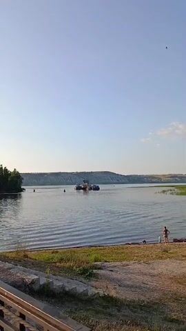 The video shows a wide river under a clear blue sky. In the distance, on the water, is a ferry with several boats. On the right bank of the river, there is a sandy area with some people. One person, a woman, is standing and facing the water. Another person, a child, is standing nearby. To the left of the river, there is a wooded area. The foreground of the video shows a paved area with some plants.