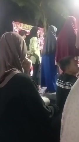 A group of people are gathered outdoors at night. In the foreground, a person wearing a black hijab and a black top is looking at their phone. To their right, a child wearing a black t-shirt with white lettering is also visible. Further back, several people are standing and sitting. One person in a light brown jacket and a checkered sarong is speaking into a microphone. Other individuals are observing, some holding cameras or phones, and one appears to be playing a flute. A banner with some red and white lettering is visible in the background, along with lights and foliage. The scene suggests an outdoor event or gathering.