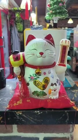 A close-up shot focuses on a ceramic Maneki-neko, a Japanese lucky cat figurine, placed on a red cushion with gold floral patterns. The cat is white with red ears and markings. It holds a gold mallet in its left paw and its right paw is raised in a waving gesture. The figurine is decorated with floral patterns and Chinese characters. A bell hangs around its neck. The background appears to be an indoor setting with a brick wall and a counter. There are other objects visible in the background, including what looks like a Christmas tree decoration and some red tassels hanging from the ceiling. The lighting is somewhat dim, casting a warm glow on the figurine. The video appears to be a short, static shot of the figurine.