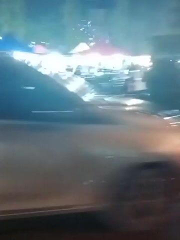 The video depicts a nighttime street scene, possibly a market or food stall area, with several vehicles and people. The camera appears to be moving, possibly from inside another vehicle, causing motion blur. Lights from stalls and vehicles illuminate the scene. One frame shows a dark-colored van or SUV with its headlights on. Another frame shows a white car. There are figures of people walking around. The ground appears wet in one section, suggesting recent rain or flooding. The overall atmosphere is of a bustling, active nighttime environment.