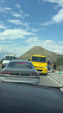 This media captures a traffic scene on a sunny day, likely in the afternoon, with a bright blue sky dotted with fluffy white clouds. The setting appears to be a roadside in a mountainous region, possibly Kruševica, Montenegro, given the rugged hills and vegetation in the background.

The main subjects are two tow trucks and a severely damaged car. The first tow truck, a bright yellow Iveco with "AUTO SLEP" written on it, is positioned in the middle of the road, presumably assisting with the accident. A man, wearing a black t-shirt, black shorts, and white sneakers, is standing near the second tow truck, a flatbed carrying the wreckage of a white car. He is looking towards the damaged vehicle and appears to be a worker involved in the cleanup or recovery operation. The damaged car is heavily compromised, with its front end completely mangled and parts scattered around.

In the foreground, the view is from inside a vehicle, showing the rear of a silver Peugeot. The overall scene suggests a recent car accident has occurred, and authorities or recovery services are on the scene dealing with the aftermath. The presence of the tow trucks and the damaged vehicle indicate a traffic disruption. Despite the unfortunate event, the weather is pleasant, suggesting the incident happened during daylight hours.