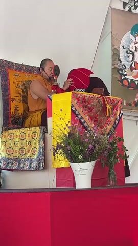 A man dressed in saffron robes sits on a cushioned, elevated seat, on a red-covered stage. He is speaking into a microphone headset and adjusting a bright red, curved headpiece that appears to be resting on his head. Behind him is an ornate textile with images in gold, yellow, and red. To his right is a table draped in bright red, yellow, and intricately patterned textiles. In front of the table is a large white bucket-shaped container overflowing with a colorful bouquet of flowers. Behind him, to the right, is a tall banner, featuring a deity and other religious motifs. The backdrop is a white tent-like structure.