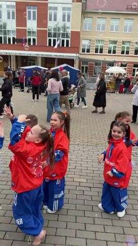The video shows a group of young children in matching red and blue tracksuits playing in an outdoor paved area. They are interacting with soap bubbles floating in the air, trying to catch them with their hands. The children are of various heights and builds, most with their hair in pigtails. They appear to be enjoying themselves, smiling and looking up at the bubbles. Behind the children, there is a crowd of adults and a few other children, some watching the bubble play. In the background, there is a multi-story brick building, a bouncy castle, and some stalls or stands that suggest an event or gathering. The sky appears overcast.