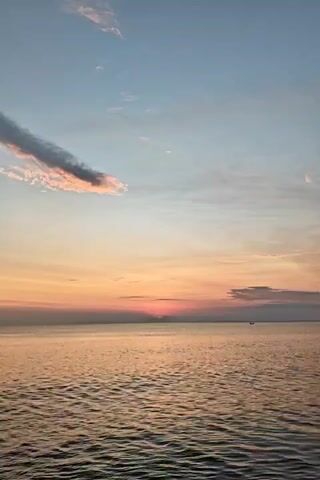 The video shows a view of the ocean at sunset. The sky is a mix of blue, orange, and pink hues, with some clouds scattered across it. The sun is setting on the horizon, casting a warm golden light on the water. The water is calm with gentle waves. There is a small boat visible in the distance.