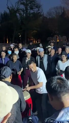 A medium shot shows a group of people gathered outdoors at dusk, listening to live music. The crowd consists mainly of older men, many wearing hats and some face masks. A man in a navy suit plays a set of two silver trumpets, while another man beats a red and black drum with two drumsticks. The environment appears to be a park or public space, with trees visible in the background and streetlights providing illumination. The atmosphere seems lively and communal as people watch and listen to the performance.