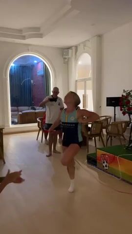 A woman in a blue and white athletic outfit and white socks is dancing in a brightly lit room with wood floors. Her hair is blonde and pulled back. Behind her, a man in a white polo shirt and dark shorts is holding up a phone, possibly recording. To her left, another person's arms are visible, clapping. In the background, there is a dining table with chairs, a large window, and what appears to be a decorative soccer-themed display. The room has white walls and a high ceiling with decorative molding. The lighting is bright and even, suggesting daytime or well-lit interior. The overall scene appears to be a casual gathering or party.