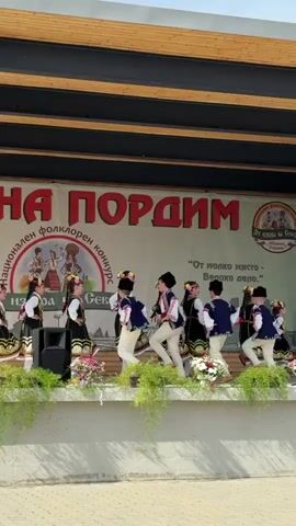 This video shows a group of young people in traditional folk costumes performing a dance on an outdoor stage. The costumes consist of black dresses with embroidered details and white accents, and the performers wear floral headpieces. Some of the boys wear vests and hats. They are arranged in rows and appear to be moving in sync. A large banner with Cyrillic text is visible behind them, along with floral decorations on the stage. The lighting suggests it is daytime.