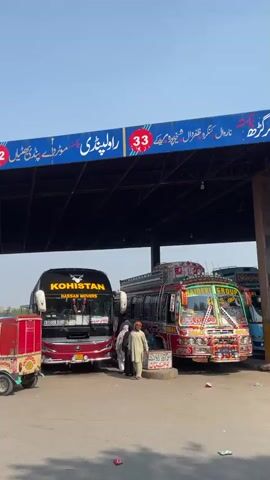 The video shows an open-air bus station in Pakistan, under a blue awning supported by posts.  The awning is numbered from 29 to 33 and has text in Urdu. Buses, auto rickshaws and cars are parked.  Several men in traditional Pakistani clothing, including shalwar kameez, are walking around the bus station. One man carries a tray on his head.  The sky is clear and blue.  The camera pans to show the full length of the bus station.