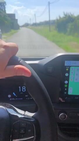 The video shows the interior of a car driving on a sunny day. The camera is positioned behind the steering wheel, looking forward through the windshield. A person's hand is visible on the steering wheel.

The dashboard features a digital speedometer displaying a speed of 19 km/h, which later increases to 22 km/h. A large central touchscreen displays a navigation app showing a map with a route. The text "SEARCH" is visible at the top of the navigation display, along with destination information: "Amazon DTH1," "46 min," and "53 km," followed by "Berliner Str. 3," "55 min," and "53 km." There is also a music app displayed on the right side of the screen, showing "HIGHLIGHTS VIP MORGAN HODGKINSON STRANDERS." The time displayed on the dashboard is 3:43.

A smartphone is mounted above the dashboard, also displaying a map with a route. The background seen through the windshield is a paved road leading through a rural area with green fields and trees on either side. The sky is blue with some white clouds. Power lines are visible in the distance. The overall impression is of a car journey in a rural setting in Germany, likely Jena given the context. The weather appears to be fair and the mood seems relaxed, indicated by the casual driving and the music playing.