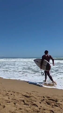 A man in a dark wetsuit walks toward the ocean, carrying a surfboard. The man is walking away from the camera on a sandy beach. The ocean is choppy with white waves. The sky is clear and blue. The beach is wide, and there are a few footprints in the sand.
