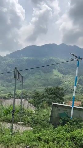This video captures a scenic journey through a rural, mountainous region in Minh Sơn Commune, Vietnam, under a cloudy, overcast sky during the daytime. The camera moves continuously, suggesting a view from a moving vehicle or person walking.

The primary subjects are the lush green mountains dominating the background, featuring varied terrain and cultivated areas that might be terraced fields. In the midground and foreground, several simple, rustic structures appear, including small houses with dark corrugated metal or tiled roofs, often made of wood. One multi-story wooden house with balconies and decorative plants is visible. Other structures include a small wooden shelter or storage area. The landscape is rich with green foliage, tall grasses, and large-leafed trees. A dirt road or path is consistently visible in the foreground, sometimes with scattered rocks, debris, or building materials. Power lines are a prominent feature, stretching across the frames throughout the video. There are no visible people or discernible text within the scene itself.