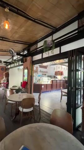 The video shows the interior of a cafe. The cafe has a rustic aesthetic, with bamboo covering the ceiling and wooden floors. There are several tables and chairs scattered around the space. In the background, there is a counter area that appears to be a display for baked goods or pastries. Plants are used as decoration, hanging from the ceiling. The lighting in the cafe is warm, with several pendant lights visible. The overall impression is of a cozy and inviting cafe environment.