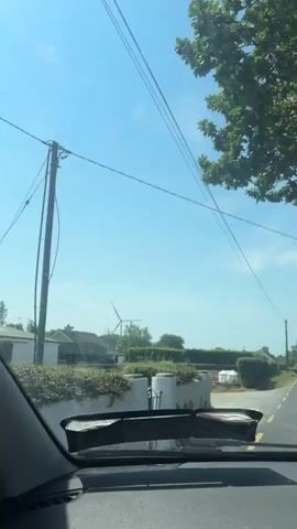 A car travels down a paved road under a clear blue sky. The windshield of the car is visible in the foreground, with a black windshield wiper assembly on top. Beyond the windshield, a landscape unfolds featuring a windmill, houses, trees, and hedges. Power lines cross diagonally across the sky. The video captures a sunny day with a hint of dust on the windshield.