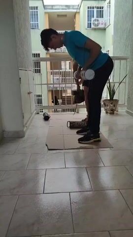The video shows a person performing a dumbbell row exercise on a balcony. The person is wearing a blue t-shirt, black pants, and sneakers. They are holding a dumbbell in each hand and bending at the waist with their back straight. The dumbbells are being lifted towards the chest. The balcony is tiled and has a railing. There is a plant in a pot to the right of the person. Another person is partially visible in the background, also appearing to be exercising.