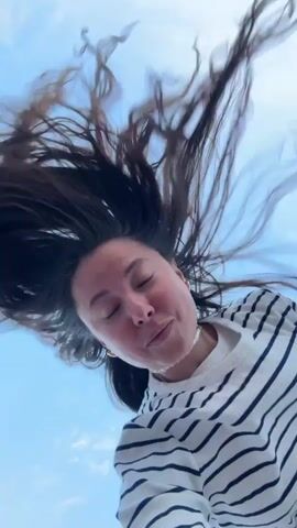 A low-angle, close-up video shows a woman with long, dark hair against a blue sky. The woman is wearing a black and white striped shirt and a pearl necklace. Her hair is flying around her head, suggesting movement, possibly from wind or her own motion. Her facial expression changes from a slight smile to a wide-eyed surprise and then to a puckered expression. The video appears to be shot from a phone being held by the woman looking up at the sky.