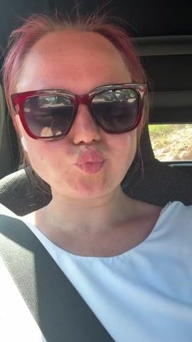 A woman with pink hair and sunglasses is in the passenger seat of a car. She is wearing a white shirt and a seatbelt. She is looking at the camera and puckering her lips. The background is blurry and appears to be a wooded area.