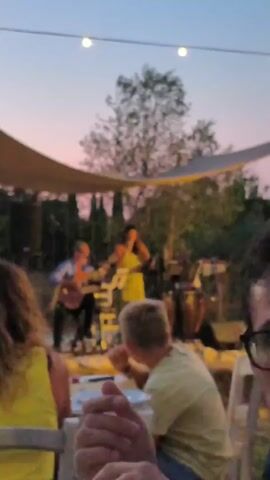 The video shows an outdoor evening event. In the background, a band performs on a stage under a canopy. The lead singer, a woman in a yellow dress, is holding a microphone and singing. To her left, a man in a blue shirt plays an acoustic guitar. To her right, another musician plays percussion, possibly congas. Several people are seated at tables in the foreground, facing the stage. A young boy in a light green shirt is among them, looking towards the stage. The sky is dusky, with string lights overhead. The overall atmosphere appears to be a casual outdoor concert or gathering.