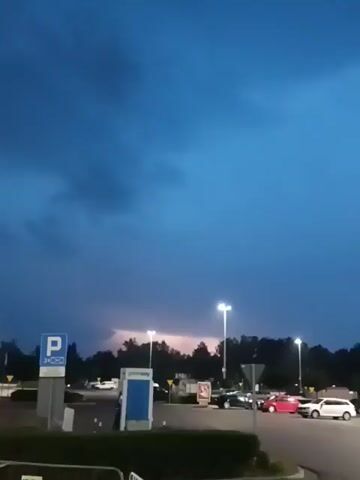 This video shows a dark blue sky with clouds. In the distance, there is a faint purple glow, possibly from lightning. In the foreground, there is a parking lot with several cars, including a white SUV, a red car, and some darker colored cars. There is also a parking sign with a blue "P" on it. Streetlights are also visible, and there are trees in the background.