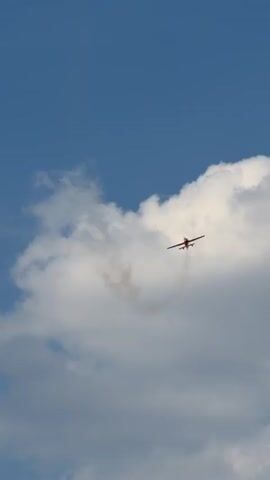 This media file contains a short video clip showing a remote-controlled airplane performing aerial maneuvers against a bright blue sky with scattered white clouds. The airplane is primarily red and white with some black accents. It is seen at various angles, sometimes leaving a white smoke trail behind it, indicating it's likely engaged in an aerobatic display. The scene suggests an outdoor event or practice session, likely during daylight hours, given the clear sky and bright lighting. The setting is an open sky, characteristic of an airfield or a park where such activities are common. There are no people visible in the frame, and the focus is solely on the airplane and the sky. The video is a brief capture of the airplane's flight, highlighting its agility and the visual effect of the smoke trails. There is no visible text within the video frames.