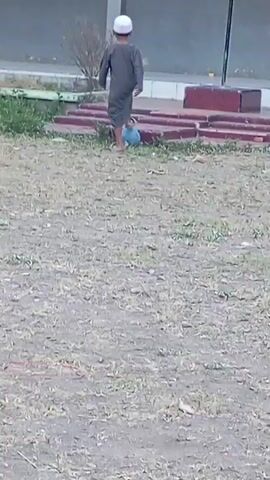 A child wearing a grey thobe and white skullcap stands in a grassy field with a blue ball in front of them. The child appears to be looking at the ball. In the background, there is a building with red benches. The scene is illuminated by natural light.