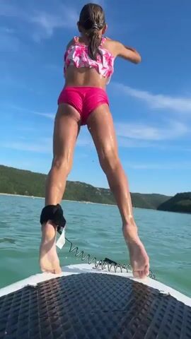A girl in a pink and white ruffled bikini jumps off a paddleboard into the water. She lands with a splash and then begins to swim. The paddleboard is in the foreground, its textured surface visible. The water is a clear, turquoise color. In the background, rolling green hills and a bright blue sky with wispy clouds are visible. The video is shot from the perspective of someone on the paddleboard, looking towards the girl.