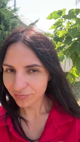 A medium close-up shot shows a woman with long dark hair and a red shirt. She is outdoors, with greenery in the background. She touches her face with her hand, which has bright blue nail polish. The camera focuses on her face as she looks at it.