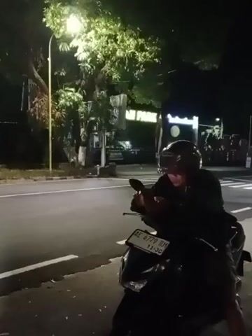 The video shows a person wearing a helmet, sitting on a motorcycle at night. The motorcycle is parked on the side of a road, next to a white line. A street light illuminates the scene, casting a glow on a tree. In the background, there are buildings and some lights. The person on the motorcycle is looking down at a phone. Another motorcycle is seen passing by in the distance, its headlights blurred due to motion. The license plate of the parked motorcycle is visible and reads "AE 6779 RM".
