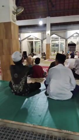 The video shows a group of people sitting on the floor inside a building. Most of the individuals appear to be men, and many are wearing skullcaps. The individuals are seated on mats or carpets. Some are facing away from the camera, while others are partially visible from the side or front. One person in the foreground, facing away from the camera, is wearing a black garment with a patterned design and a white skullcap. They are holding up a mobile phone, suggesting they might be recording or taking pictures. The environment appears to be a communal space, possibly a mosque or a similar religious or gathering hall, indicated by the presence of pillars, arched doorways in the background, and what appear to be prayer mats. The lighting suggests it might be daytime or an indoor space with artificial lighting. The overall scene depicts a group of people gathered in a calm and seated posture.