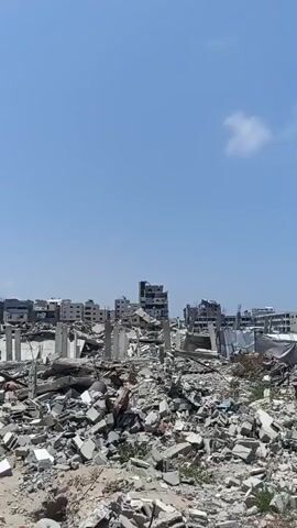 The video depicts a scene of widespread destruction in Jabalia, Palestinian Territories. The foreground is dominated by piles of rubble, concrete debris, and twisted metal, remnants of destroyed buildings. Standing concrete pillars, some intact and others broken, punctuate the landscape of devastation.

In the background, partially destroyed multi-story buildings are visible, with some appearing to be completely gutted or collapsed. The architecture suggests a residential or urban area that has suffered significant damage. The overall impression is one of a post-conflict or disaster zone, with buildings reduced to ruins.

The sky above is clear and blue, with a few scattered white clouds, indicating a daytime setting, likely mid-morning or afternoon. The weather appears to be sunny and clear. There are no people visibly present in the frame. The scene evokes a sense of loss and devastation. No visible text is present in the video.