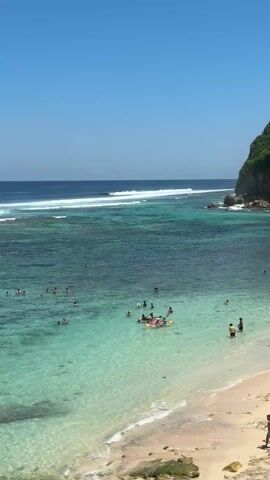 The video shows a beautiful beach on a sunny day. The water is clear and turquoise, with a few waves breaking in the distance. Many people are swimming and enjoying the water. Some are on an inflatable raft in the middle of the bay. A man walks along the sandy beach in the foreground. The beach is nestled in a cove, and a rocky cliff covered in green vegetation rises on one side. The sky is clear and blue.