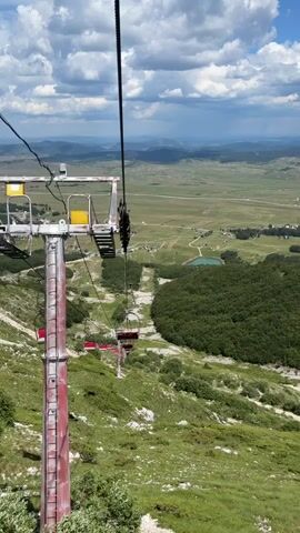 The video shows a view from a ski lift ascending a mountain on a cloudy day. In the foreground, a red and white ski lift tower with a metal staircase is visible. A cable runs from the top of the tower, with several red ski lift chairs attached to it. The chairs are moving away from the viewer, down the slope. Below the ski lift, the mountain is covered in green grass and scattered bushes. In the middle ground, a dense forest of dark green trees forms a curved shape. Beyond the forest, a wide, green valley stretches out, dotted with buildings and a small blue lake. In the background, rolling hills and mountains fade into the distance under a sky with scattered white clouds and patches of blue. The overall scene depicts a mountain landscape in warmer weather, likely during the summer or early autumn, given the greenery.