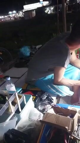 A man in a gray t-shirt and blue shorts sits cross-legged on the ground outdoors at night, working on a small electronic device. He is under the light of a white and red lamp. His work area consists of a blue tray covered with tools such as pliers, screwdrivers, and small containers. There's a power strip plugged in nearby, and various boxes, containers, and a water bottle are scattered around. In the background, a street scene with shops and passing vehicles is visible. The man focuses intently on the device in his hands, seemingly repairing or modifying it.