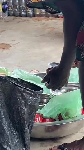The camera records the scene of someone preparing food or gathering supplies. An individual with dark skin, wearing a green garment, is reaching into a large metal bowl. This bowl is filled with items in light green plastic bags, alongside small bowls, and some red rectangular items. A black plastic trash bag is partially visible on the left side of the frame. In the background, there is an assortment of bottled drinks, and the floor appears to be concrete with some stains or marks on it. The person's action suggests they are organizing or packing items.